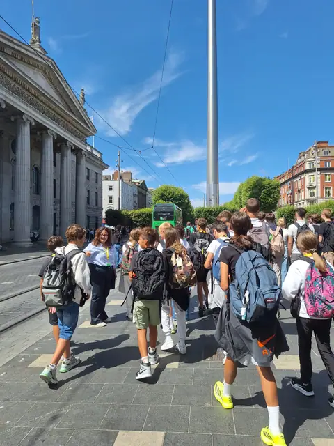 Il gruppo cammina lungo un ampio viale del centro, tra un palazzo con colonnato e un’alta guglia d’acciaio