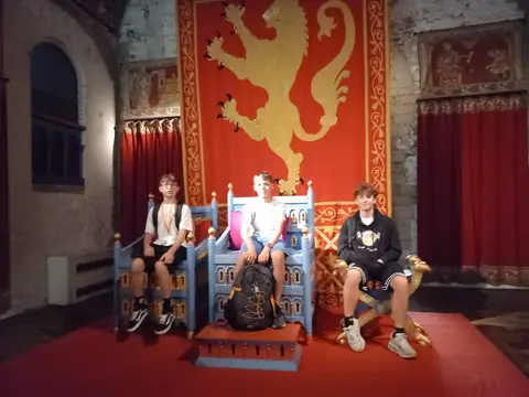 Tre ragazzi seduti su troni in una sala medievale con un arazzo rosso e un leone dorato