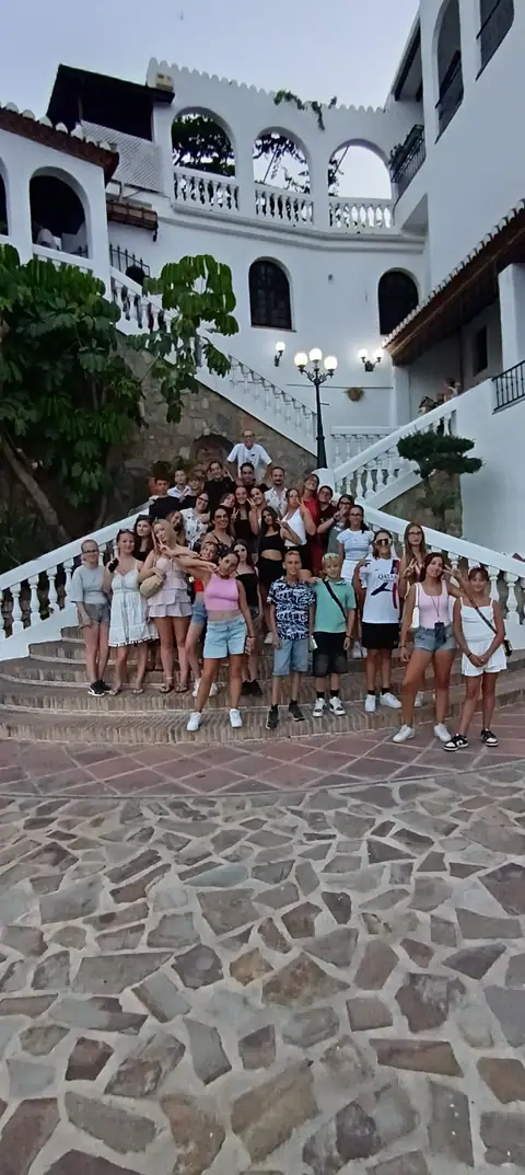 Un gruppo di ragazzi in posa sulla scalinata di un edificio bianco con archi e balconi, tra palme e lampioni